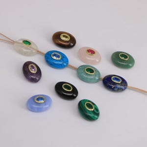 Puede incluir: Una colecci&oacute;n de cuentas de piedras preciosas ovaladas en varios colores, incluyendo azul, verde, rosa y negro. Cada cuenta presenta un engaste dorado y una peque&ntilde;a gema de colores en el centro. Las cuentas est&aacute;n ensartadas en un fino cord&oacute;n beige.