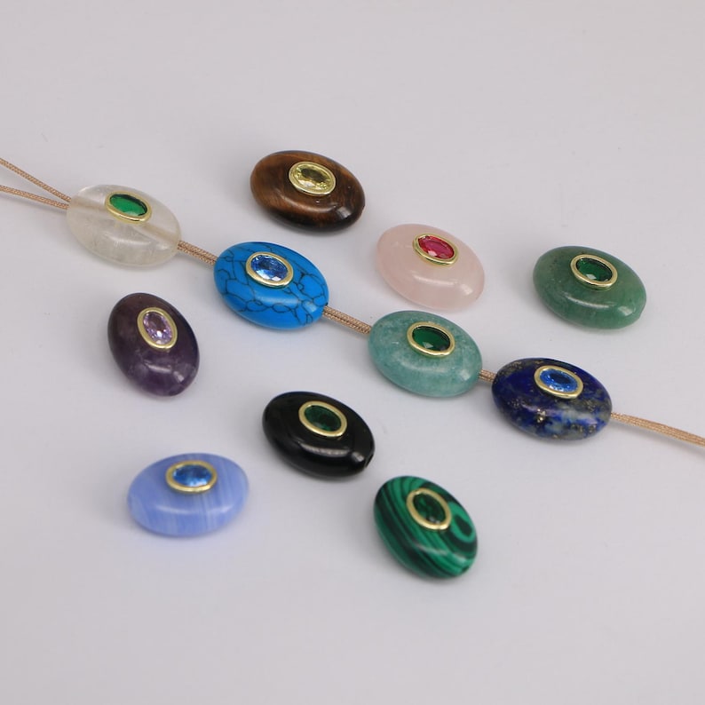 Puede incluir: Una colecci&oacute;n de cuentas de piedras preciosas ovaladas en varios colores, incluyendo azul, verde, rosa y negro. Cada cuenta presenta un engaste dorado y una peque&ntilde;a gema de colores en el centro. Las cuentas est&aacute;n ensartadas en un fino cord&oacute;n beige.