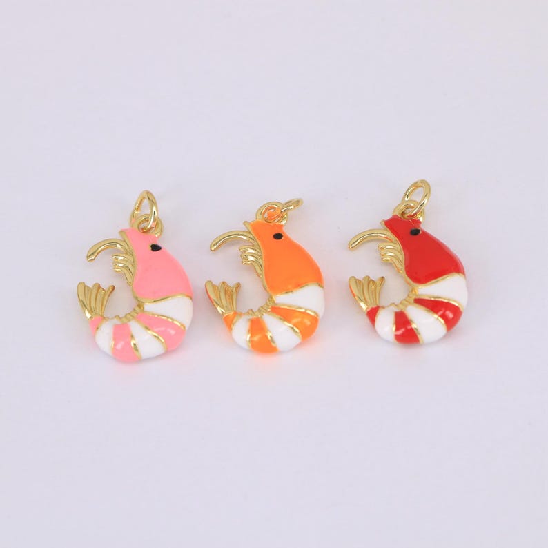 May include: Three colorful shrimp charms in pink, orange, and red with white stripes. Each charm has a gold-colored loop for attaching to jewelry. The shrimp are stylized with a curved body and detailed tail.