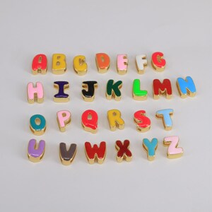 May include: A collection of alphabet beads in a variety of colours, including red, yellow, green, and blue. Each letter has a gold-coloured border. The letters are arranged in rows, displaying the entire alphabet.