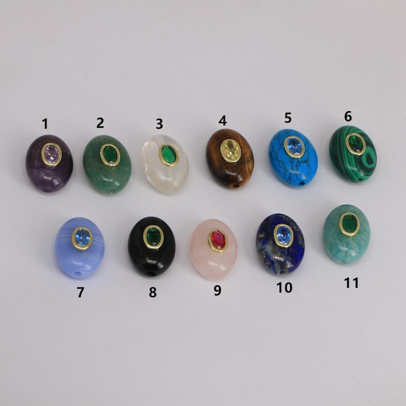 Puede incluir: Once cuentas de piedras preciosas ovaladas en varios colores, incluyendo p&uacute;rpura, verde, transparente, marr&oacute;n, turquesa y verde azulado. Cada cuenta presenta una piedra preciosa m&aacute;s peque&ntilde;a y contrastante engastada en un bisel dorado. Las cuentas est&aacute;n dispuestas sobre una superficie blanca.