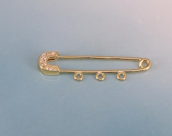 May include: A gold-colored safety pin with a curved top encrusted with small, clear stones. The pin has three small, circular loops along the straight edge. The background is a gradient of light blue and white.