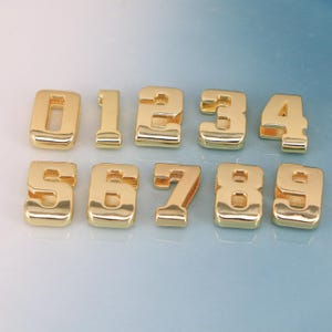 May include: A collection of ten gold-colored metal number charms, ranging from zero to nine. Each number has a rounded, blocky font with a polished finish. The charms are arranged in two rows on a light blue background.
