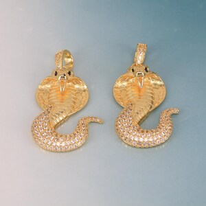 Pode incluir: Dois pingentes de cobra dourados. As cabeças de cobra apresentam escamas texturizadas e olhos pretos. O corpo da cobra é curvo, incrustado com pequenas pedras preciosas transparentes. Os pingentes têm uma argola na parte superior para uma corrente.