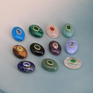 Puede incluir: Una colecci&oacute;n de cuentas de piedras preciosas de forma ovalada en varios colores, incluyendo azul, verde, rosa y morado. Cada cuenta presenta un engaste dorado alrededor de una piedra preciosa m&aacute;s peque&ntilde;a y contrastante. Las cuentas est&aacute;n dispuestas sobre una superficie azul claro.