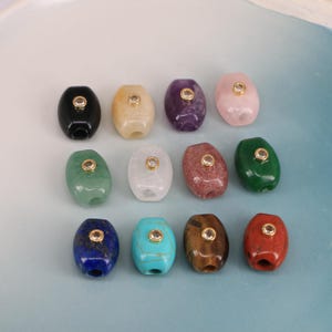 May include: An assortment of oval-shaped gemstone beads in various colors, including black, yellow, purple, pink, green, white, red, blue, turquoise, and brown. Each bead has a gold-colored metal accent in the center. These beads are suitable for jewelry making.