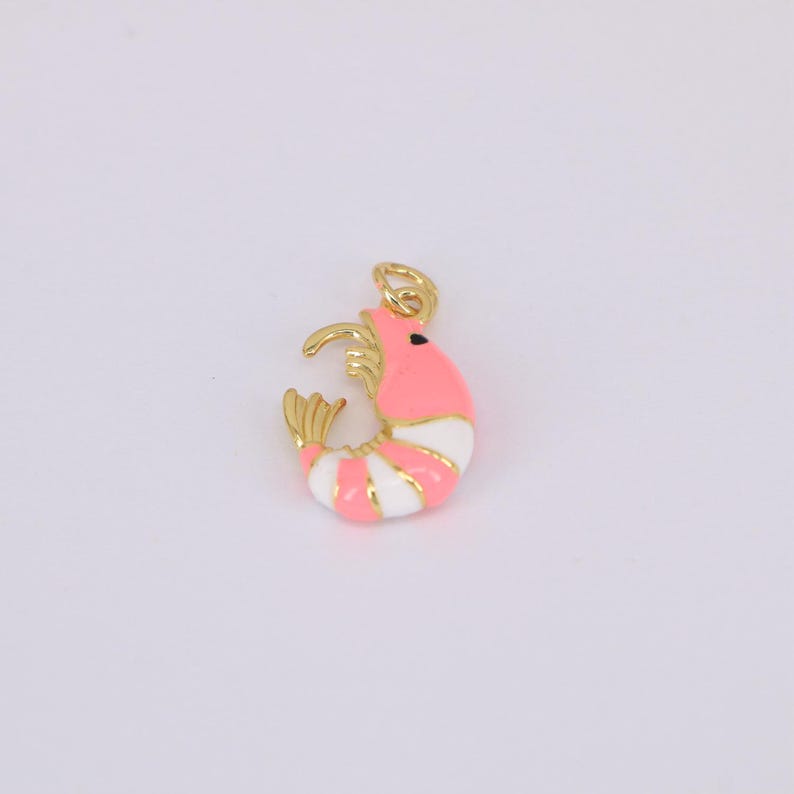 May include: A gold-colored metal shrimp charm with a pink body, white stripes, and a gold-colored tail and head. The charm has a small loop at the top for attaching to a necklace or bracelet. The shrimp's eye is a small black dot.