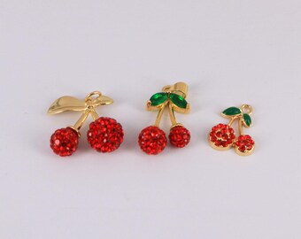 May include: Three gold-toned cherry charms with red crystal-like spheres and green leaf accents. The charms vary in size and design, with one featuring a bow-like stem and another with a more detailed leaf structure. The cherries are a vibrant red.