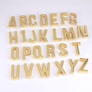 May include: A complete set of twenty-six individual uppercase letters of the alphabet, crafted from a shiny gold-toned metal. Each letter features a smooth, reflective surface and a three-dimensional block font design, arranged in rows.