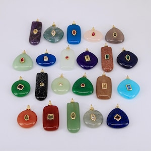 Puede incluir: Una colección de colgantes de piedras preciosas en varias formas y colores, incluyendo púrpura, verde, azul y marrón. Cada colgante presenta una fianza dorada y una pequeña gema incrustada. Los colgantes están dispuestos sobre una superficie blanca.