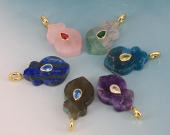 Puede incluir: Una colección de colgantes de piedras preciosas en varios colores, incluyendo rosa, verde, azul y morado. Cada colgante presenta una fianza dorada y un acento de piedra preciosa en forma de lágrima. Los colgantes están dispuestos sobre una superficie azul claro.