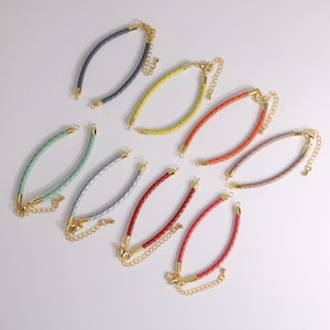 May include: A collection of colorful braided leather bracelets with gold-tone clasps and extender chains. The bracelets are in various colors, including gray, yellow, orange, pink, red, and green. The bracelets are displayed on a white surface.