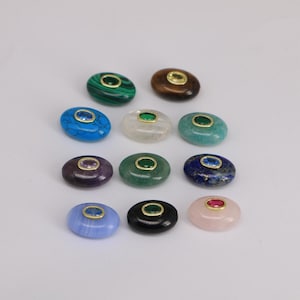 Puede incluir: Un surtido de piedras preciosas ovaladas en varios colores, incluyendo azul, verde, morado y rosa. Cada piedra est&aacute; engastada en un bisel dorado y presenta una piedra preciosa m&aacute;s peque&ntilde;a en el centro. Las piedras preciosas est&aacute;n dispuestas sobre una superficie blanca.