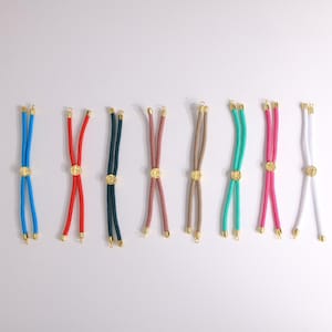 May include: A selection of colourful rope necklaces with gold-coloured accents. The necklaces are in various colours, including blue, red, green, pink, and white. Each necklace features a circular gold-coloured charm and gold-coloured end caps.