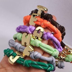 Puede incluir: Una colección de pulseras de cuerda coloridas, cada una con un dije de letra dorada. Las pulseras son en tonos de verde, gris, morado, naranja y marrón. Las letras son en negrita, sin serifa.
