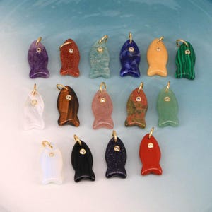 Puede incluir: Una colección de colgantes de pez de piedras preciosas en varios colores, incluyendo morado, rojo, verde y azul. Cada colgante tiene un lazo dorado y un pequeño acento redondo. Los colgantes están dispuestos sobre una superficie azul claro.