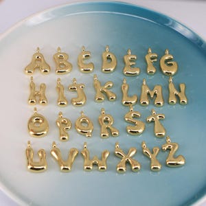 May include: A collection of gold-colored alphabet letter charms, each with a loop for attaching to jewelry. The letters are in a rounded, bubble-like font, displayed on a light blue and white plate.