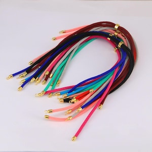 May include: A collection of colorful braided cords with gold-colored metal end caps. The cords are in various colors, including pink, blue, black, red, and green. The cords are arranged in a curved shape against a white background.