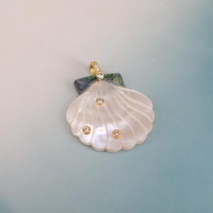 May include: A scallop shell pendant crafted from iridescent white mother-of-pearl with a dark green abalone shell accent at the top. The pendant features four small, round, clear stones set in gold-toned metal and a gold-toned bail at the top.