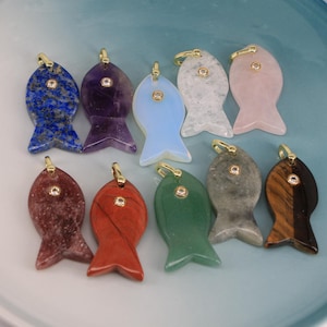May include: A collection of ten fish-shaped pendants in various colors and materials. The pendants include blue lapis lazuli, purple amethyst, and pink rose quartz. Each pendant has a gold-colored bail and a small, clear gem.