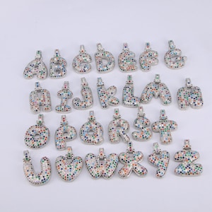 Op de afbeelding: Een set van 26 zilveren hangers in de vorm van letters van het alfabet, elke letter is bedekt met kleurrijke edelstenen.