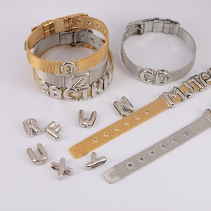 Könnte beinhalten: Gold- und silberfarbene Mesh-Armbänder mit Metallbuchstaben-Anhängern, die Namen buchstabieren. Einzelne Metallbuchstaben und zusätzliche Mesh-Armbandbänder werden ebenfalls angezeigt. Die Armbänder haben einen Schnallenverschluss.