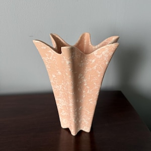 May include: A peach-colored ceramic vase with a star-shaped rim and a white speckled pattern across its surface. The vase features a flared, multi-pointed opening and a tapered body that narrows toward the base, creating a distinct star silhouette.