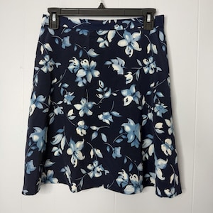 May include: A navy blue skirt with a floral pattern featuring light blue and white flowers and leaves. The skirt is hanging on a black hanger against a white background. The skirt has a slight A-line shape.