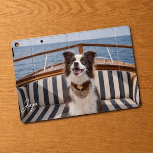 May include: A rectangular tablet case featuring a printed image of a brown and white dog wearing a patterned bandana and a gold chain, sitting on a striped bench on a boat deck. The word Jaja is written in white cursive script on the lower left.