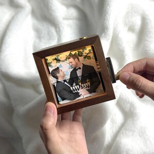 May include: A small, square wooden music box featuring a photograph of two men in formal attire. The box is held in a person's hands, with a crank on the side. The photograph shows the men in a romantic setting, with champagne glasses visible.