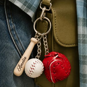 Könnte beinhalten: Ein Metallschlüsselanhänger mit drei Anhängern: einem hellen Holz-Baseballschläger mit der Gravur Amelia, einem weißen Baseball mit roten Nähten und einem kleinen roten Baseballhandschuh aus Leder mit sichtbaren Nähten, befestigt an einem Ring.