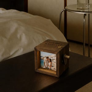 Peut inclure: Un cadre photo cubique en bois brun foncé présentant une photographie carrée d'un couple debout sur une plage avec des bâtiments blancs en arrière-plan. Le dessus du cadre en bois est gravé du texte Together wherever et possède une petite épingle dorée.