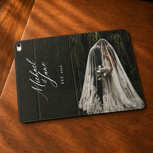 May include: A black tablet case featuring a printed wedding photograph of a couple under a veil on the right side. The left side displays the white script text Michael & Jane and the text EST. 2026. The case has a camera cutout at the top left.
