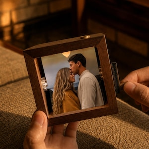 May include: A square wooden frame containing a photograph of a person with long blonde hair wearing a mustard yellow top and a person in a light-colored shirt leaning in close. The frame features a small metal crank handle on the right side.