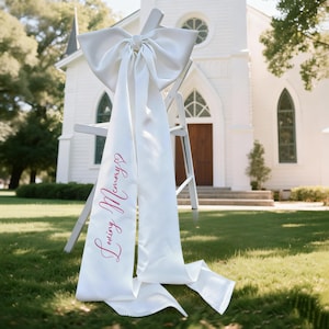 Op de afbeelding: Witte satijnen stoelsjerp met een grote strik en de woorden "Loving Memory" in roze script. De sjerp is over een witte stoel gedrapeerd, met een kerk op de achtergrond. De strik is bovenaan de stoel vastgebonden.