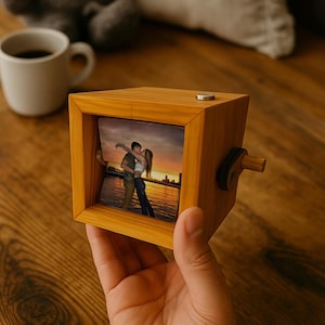 Puede incluir: Un cubo de fotos de madera con una foto de una pareja besándose. El cubo tiene un acabado marrón claro y una pequeña manivela en el lateral. Una taza blanca de café está en el fondo.