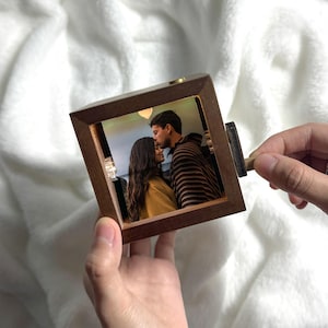 May include: A wooden photo cube displaying a photograph of a couple. The cube has a dark brown frame and a small wooden handle. The photo is lit by a soft light. The cube is held in a person's hand.