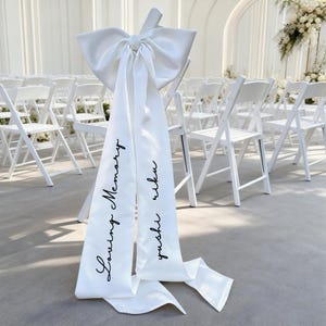 Puede incluir: Un lazo de silla de satén blanco con largas cintas fluidas. Las cintas están impresas con las palabras "Loving Memory" y "yushi riku" en escritura negra. El lazo está atado a una silla plegable blanca, con filas de sillas similares en el fondo.