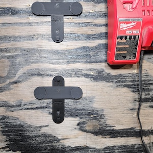May include: Two black, T-shaped mounting brackets with circular ends are mounted on a wooden surface. The text "This side up" and "Milwaukee Charger Mount" are visible on the brackets. A red Milwaukee M12/M18 charger is also present.