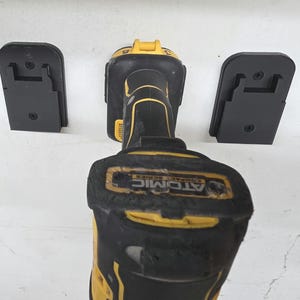 Op de afbeelding: Zwarte en gele boormachine met twee zwarte montagebeugels. Op de boormachine staat de tekst "ATOMIC COMPACT SERIES". De beugels zijn ontworpen om de boormachine vast te houden.