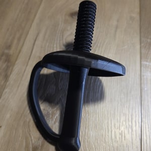 May include: A black, 3D-printed sword hilt featuring a circular guard, a cylindrical handle with a rounded pommel, and a threaded bolt extending from the top of the guard. The object is constructed from a matte black material with visible layer lines.