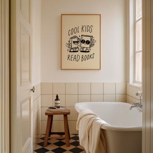 K&ouml;nnte beinhalten: Eine Schwarzwei&szlig;illustration von zwei B&uuml;chern, die Sonnenbrillen tragen und gehen, mit dem Text "Cool Kids Read Books" dar&uuml;ber. Die Illustration ist gerahmt und h&auml;ngt an einer Wand in einem Badezimmer mit einer wei&szlig;en Badewanne und einem Holzhocker.