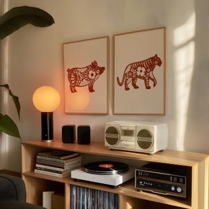 Puede incluir: Dos impresiones enmarcadas de ilustraciones de animales rojas y blancas con motivos florales. Las impresiones est&aacute;n colgadas en una pared sobre un estante con una l&aacute;mpara, altavoces, un tocadiscos y un sistema est&eacute;reo.