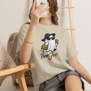 May include: A beige short-sleeve t-shirt featuring a graphic of a ghost wearing a black hat, sunglasses, and holding a coffee cup and a leopard print handbag. The text "BooJee" is printed below the ghost in a stylised black and leopard print font.