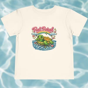 Puede incluir: Una camiseta de manga corta color crema con un gráfico de una tortuga verde en un flotador amarillo sobre agua azul. Sobre la tortuga hay un arcoíris, nubes y el texto Pool Patrol en letras rosas, rodeado de pequeños corazones y estrellas.