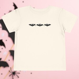 May include: A cream-colored short-sleeve t-shirt featuring a horizontal row of three black bat illustrations centered on the chest. Each bat is depicted with small star accents between them, creating a simple graphic design on the plain fabric.