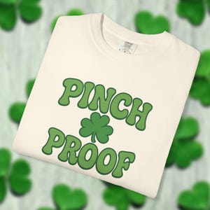 Puede incluir: Camiseta color crema con las palabras "PINCH PROOF" en verde y un trébol verde. La camiseta está doblada y se muestra sobre un fondo con formas de tréboles verdes.