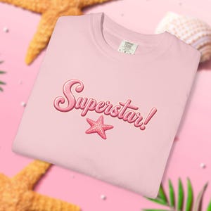 Puede incluir: Una camiseta de color rosa claro con la palabra Superstar! en una fuente brillante de estilo burbuja y un gráfico de una estrella de mar rosa situado justo debajo del texto. La prenda tiene cuello redondo y una etiqueta de marca visible.