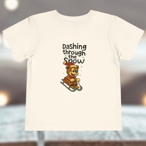 Puede incluir: Una camiseta de manga corta de color crema con un gráfico de un oso marrón con gorro y pajarita rojos, montado en un trineo. Sobre el oso, el texto Dashing through the Snow aparece en letras de color marrón oscuro.