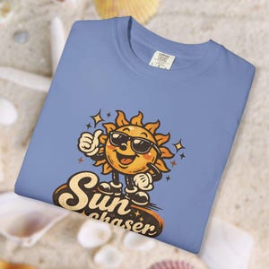 Puede incluir: Camiseta azul claro con un sol de dibujos animados con gafas de sol y un pulgar hacia arriba. El texto "Sun Chaser" está debajo. La camiseta es de material suave.